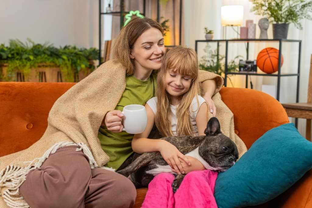 Parent and child wrapped in a blanket on a sofa with a small dog, looking warm and comfortable