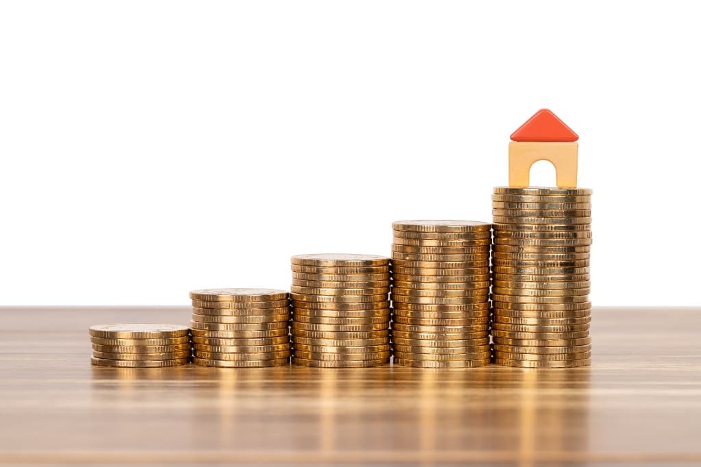Stacks of gold coins rising like steps with a small wooden toy house on the tallest pile